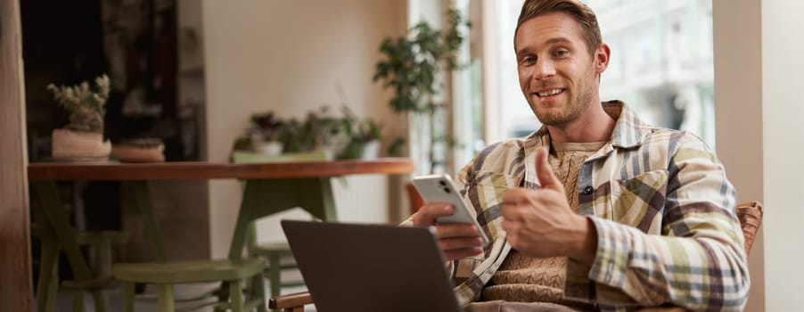 Portrait of smiling, handsome young man relaxing in coffee shop, working remotely from cafe co-working space, using laptop and smartphone, looking at camera and showing thumbs up with satisfied face.