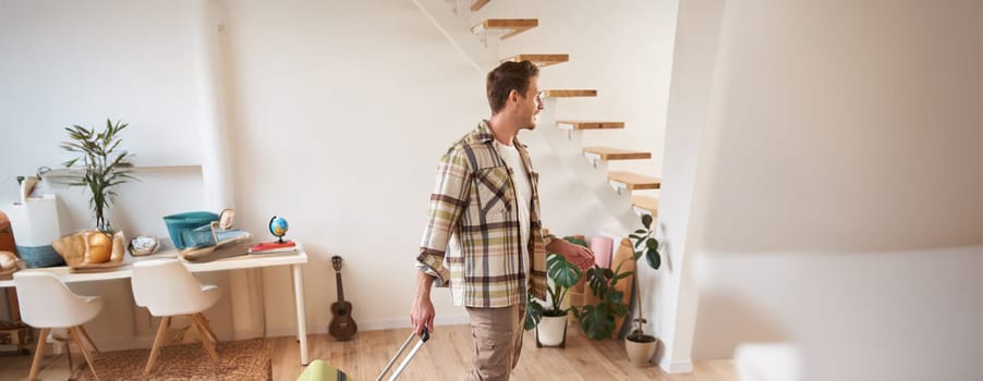 Vertical portrait of young handsome man walking indoors with suitcase, going on vacation, leaving his rented apartment with luggage.