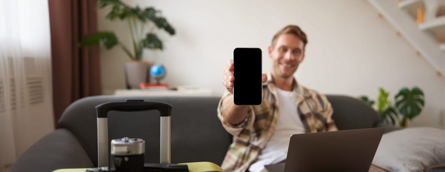 Portrait of handsome young man, showing smartphone screen, recommending mobile app for travelling or booking online, sitting with laptop and suitcase. Copy space