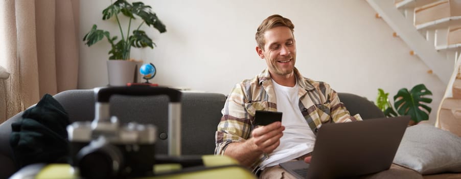 Smiling handsome guy with credit card and laptop, tourist booking a hotel for holiday, sits near suitcase.