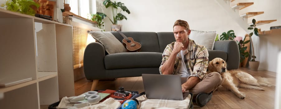 Image of young man, tourist going on holiday, sitting with a dog in living room, looking at laptop, check-in on a flight on computer. Tourism and travelling concept