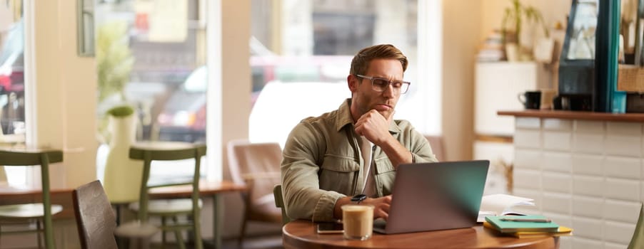 Portrait of businessman in glasses, sits in cafe with laptop, looks concentrated at screen with his project task, works remotely from coffee shop.