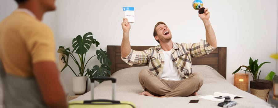 Image of happy smiling man, cheerful tourist sits with globe and plane tickets, goes on holiday, sits with suitcase, excited about his trip, going on holiday. Tourism and travel agency concept