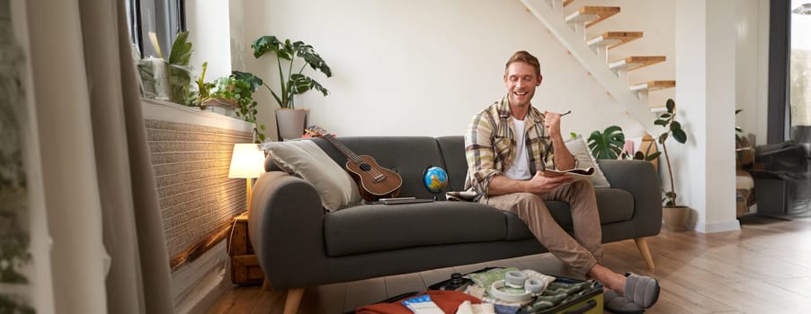 Happy man with pen and notebook, checking list of items and packing suitcase for journey, going on holiday, preparing for summer vacation, sitting in living room.
