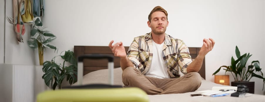 Close up portrait of meditating tourist, man sits with suitcase in bedroom, practice yoga, relaxing, finding nirvana. Tourism and travelling concept