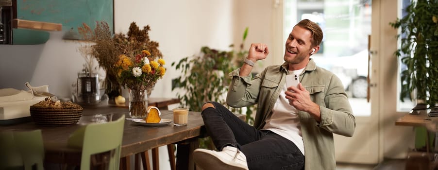 Portrait of handsome happy man in cafe, listens to music in wireless earphones, holding smartphone, connects to public wifi and enjoys favourite song, relaxing in coffee shop.