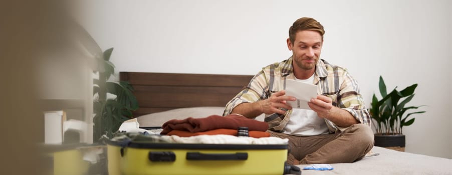 Portrait of handsome man, tourist, looking at his holiday tickets, booked vacation with travel agency, sitting on bed and packing clothes in suitcase.