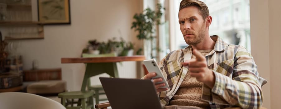 Portrait of man with serious face, sitting in cafe with laptop and smartphone, pointing at you with warning, scolding you, frowning with disapproval.