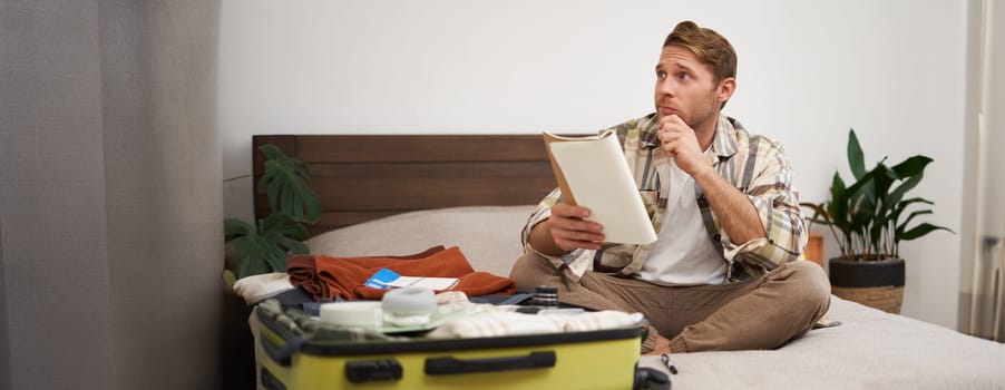 Portrait of thoughtful young man, tourist planning his vacation, sitting with open suitcase on bed, making notes, writing to do check list in notebook.