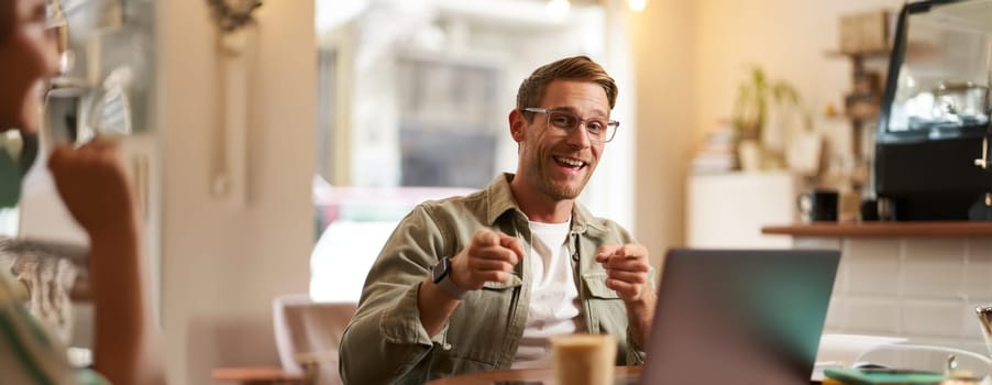 Portrait of cheerful guy in glasses, sits in cafe and works on laptop, pointing fingers at camera, inviting you, recommends you something, promoting product or public space.