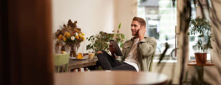 Portrait of handsome smiling young man, sitting in coffee shop, using digital tablet, video chats with someone from a cafe, wearing wireless headphones.