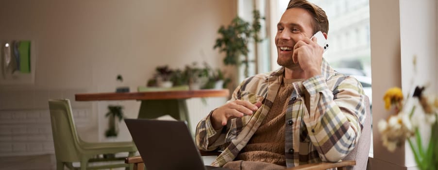 Lifestyle shot of young happy man sitting in cafe with laptop, answer phone call, talking to friend on mobile and laughing, chatting to someone with joyful face, resting in chair.