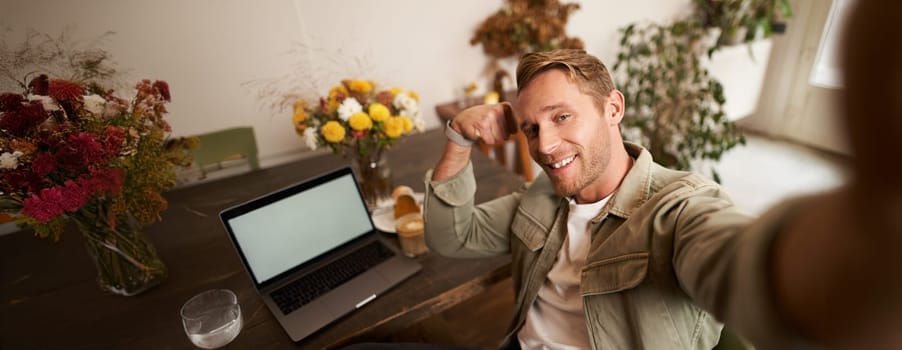 Portrait of handsome and confident man taking a selfie, flexing biceps, shows-off his muscles, sits in cafe in front of laptop, smiling at camera.