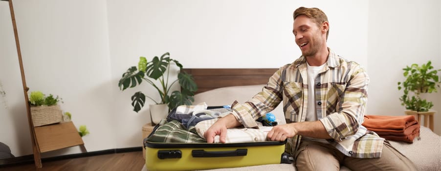Portrait of cheerful young man preparing for summer holiday, going on vacation, packing his suitcase with clothes and smiling excited. Lifestyle, people and travelling concept