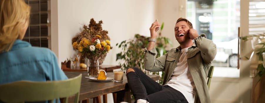 Lifestyle portrait of handsome young man, sitting in cafe, talking to a friend on mobile phone with wireless earphones, laughing and smiling, listening to funny podcast.