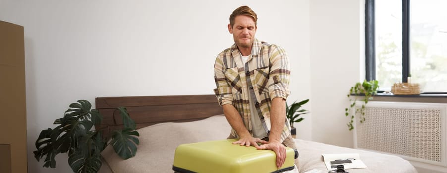Portrait of young man, tourist packs his bag, closing full suitcase, trying to push the luggage inside to zip the bag, sitting on bed in bedroom. Tourism and adventure concept