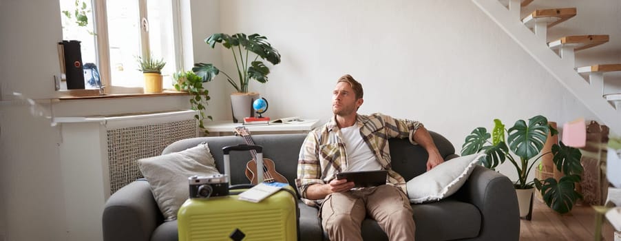 Image of young man with digital tablet, tourist sitting on sofa with suitcase packed and ukulele, thinking, looking aside thoughtful, planning holiday trip. Tourism concept