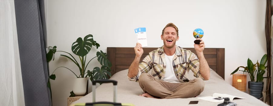 Portrait of cheerful, happy tourist, man sitting with plane tickets and a globe, packed suitcase for travelling around the world, going on vacation, excited about his holiday.