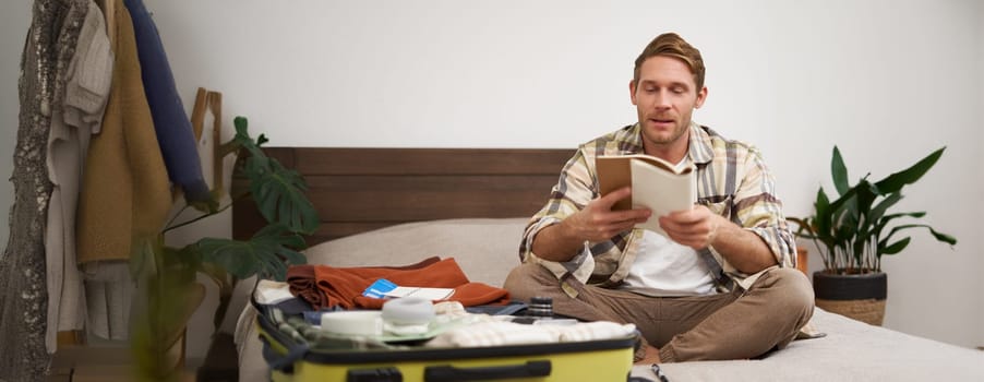 Portrait of man with suitcase, sitting and packing things for holiday, checking list of items for vacation, writing down, making notes in notebook. Lifestyle and tourism concept
