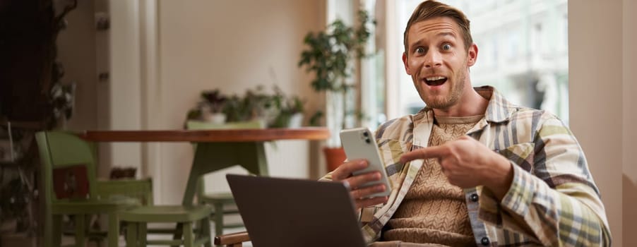 Excited guy in cafe, sitting with phone and pointing at screen with interest, recommending an app, using mobile device and laptop in co-working space. concept of freelance and remote workspace.