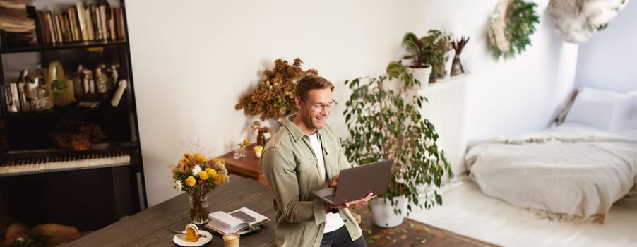 Vertical portrait of handsome happy man, manager or CEO of company, sitting on table and holding laptop, laughing, video chatting, has an online meeting with a team, standing in an office.