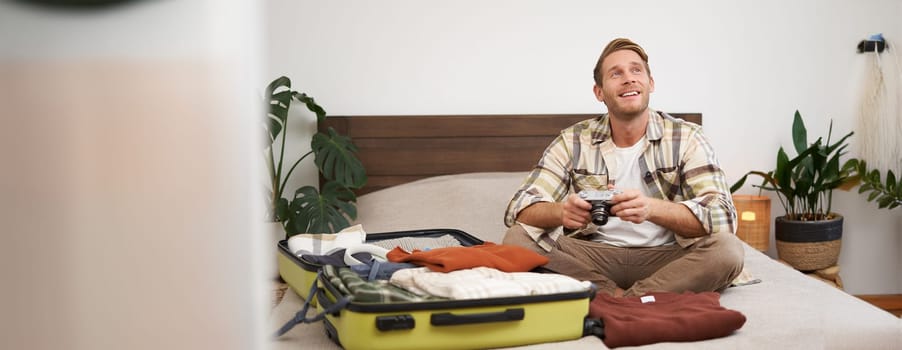 Portrait of young excited man, sits on bed with suitcase, going on holiday, holding digital camera, going through photo album on gadget, preparing for summer vacation.