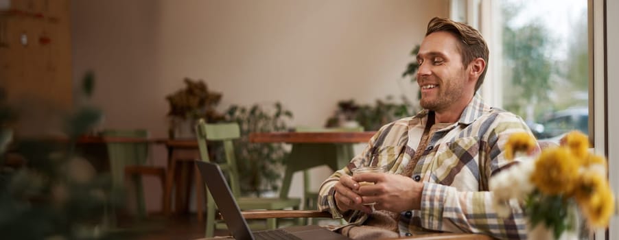 Image of handsome businessman working from cafe, drinking coffee and doing online project on his laptop, smiling and looking happy, finished his freelance task, looking pleased.