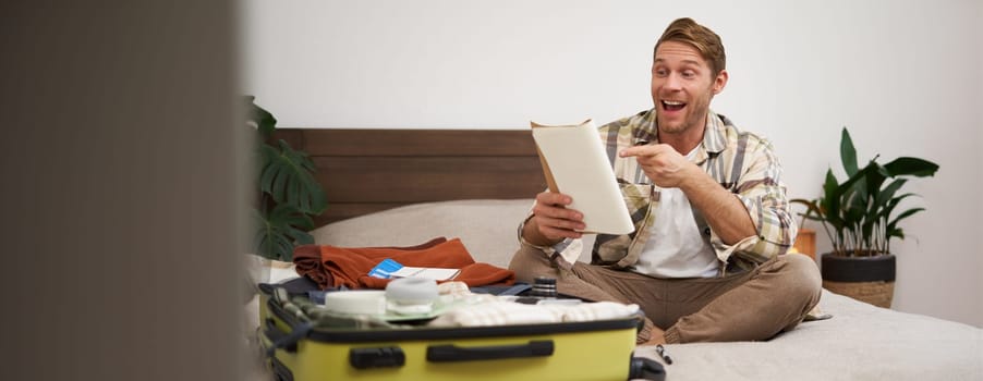 Portrait of charismatic, handsome man, tourist looking at notebook, getting ready for journey, packing suitcase, checking list of items on journal.