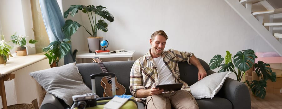 Image of young man, tourist goes on holiday, packed suitcase, sits with digital tablet on sofa, reads, looks at his map, books hotel for business trip. Copy space