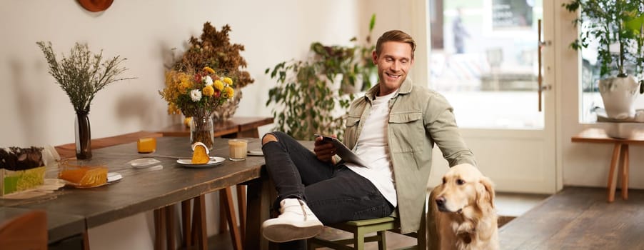 Portrait of happy smiling young man, cafe visitor, sitting in coffee shop with his dog, petting golden retriever, holding digital tablet, reading news or browsing on internet.