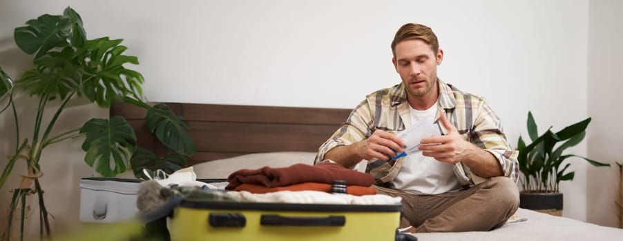 Portrait of handsome man, tourist, looking at his holiday tickets, booked vacation with travel agency, sitting on bed and packing clothes in suitcase.