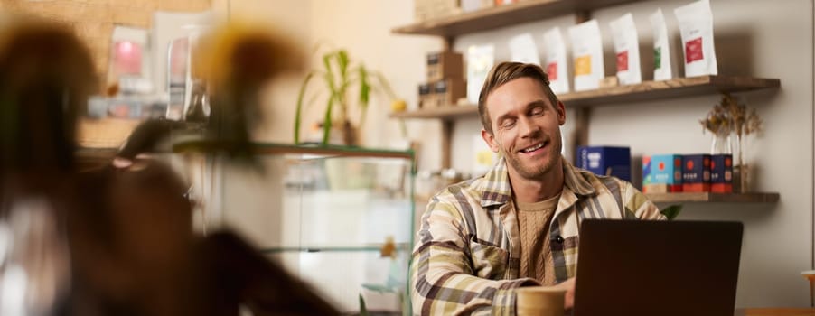 Portrait of handsome young digital nomad, man working in cafe on laptop, looking happy and pleased with his online project, sitting in co-working coffee shop.