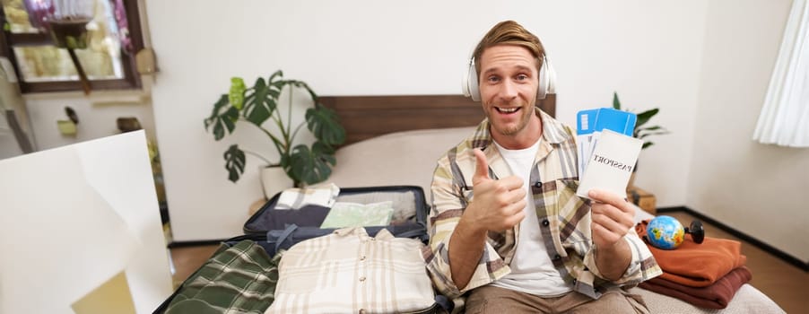 Portrait of smiling man, tourist shows two tickets and passport, goes on holiday, traveller makes thumb up in approval, like travel agency. Copy space