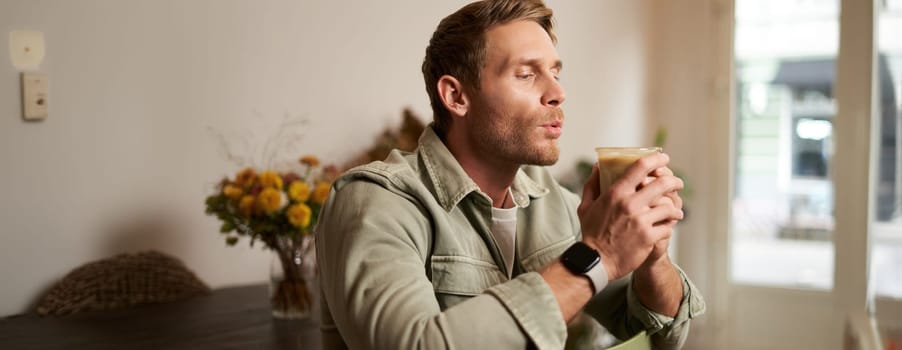 Portrait of young handsome bearded man in coffee shop, sitting on chair, sipping cappuccino, resting in cafe.