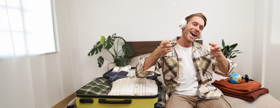 Handsome blond man in headphones, sits on bed with suitcase and clothes, listens to music in wireless headphones while packing for a holiday, going on trip.