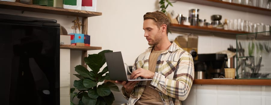 Image of young stylish guy with laptop, sitting in cafe with his computer, typing something, drinking coffee and doing project.