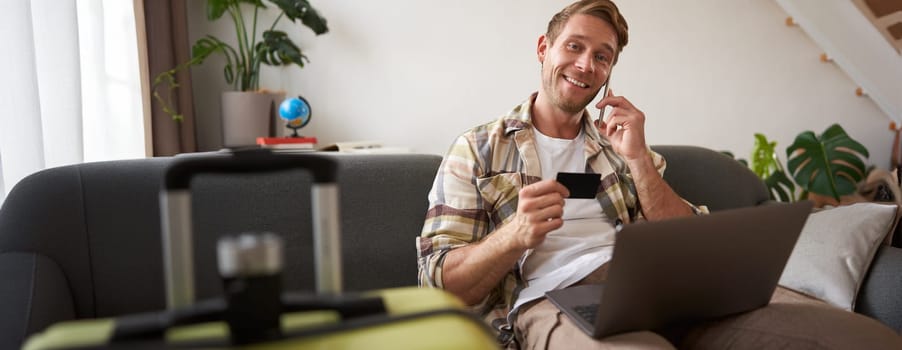 Portrait of young man with credit card, sitting and using laptop, making a phone call, booking hotel or tickets, confirming his purchase over telephone, has suitcase near him.