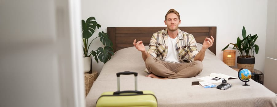 Portrait of young man meditating on a bed after packing suitcase for summer holiday, sitting with crossed legs in zen, nirvana pose, relaxing, hoping for something. Tourism and mindfulness concept
