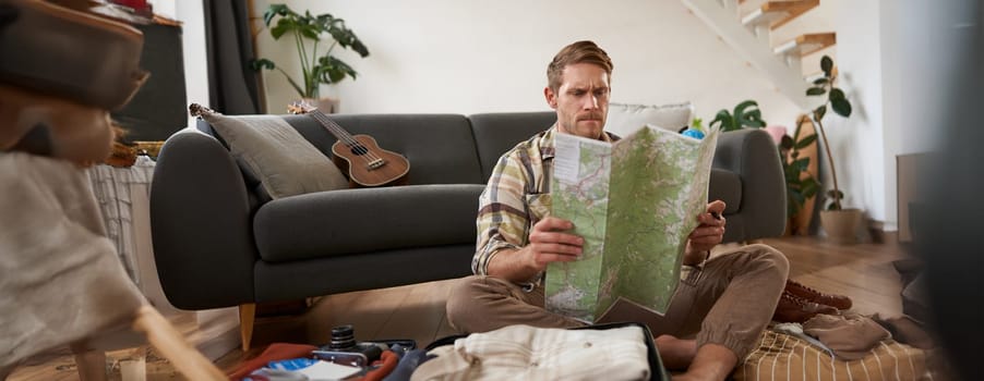 Portrait of young man, traveller, sitting packing suitcase, looking at travel map, choosing holiday destination, studying the route on vacation. Tourism concept