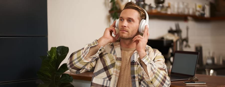 Portrait of handsome happy guy, freelancer working and listening to music in wireless headphones, dancing on his chair in cafe, enjoying favourite song in earphones.
