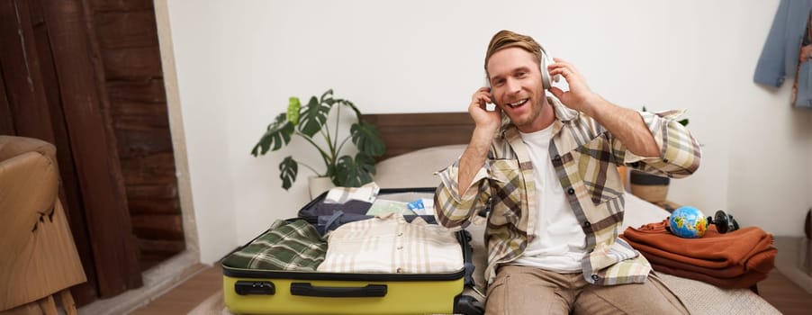Portrait of young happy man, listens to music in headphones, sits with suitcase in hotel room, unpacks clothes on vacation, tourist goes on holiday. Lifestyle and people concept