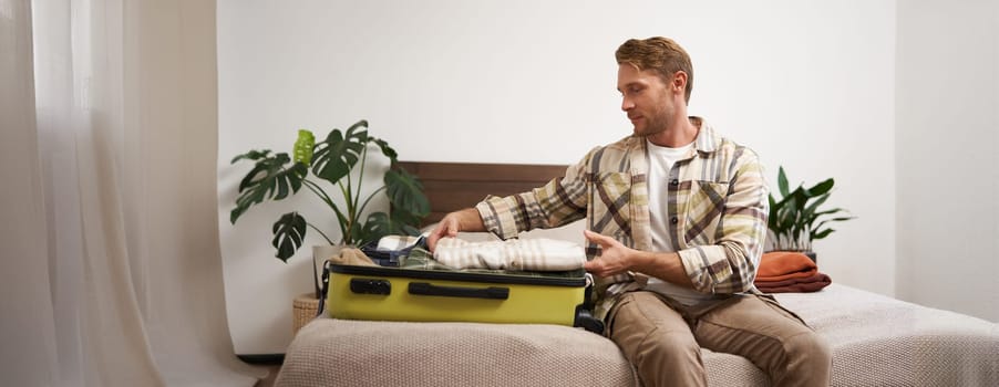Portrait of young man packing his luggage for a trip, going on holiday, sitting on bed and putting clothes in suitcase, preparing for his flight.