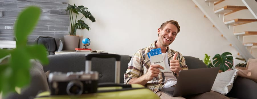 Portrait of happy handsome guy, shows his flight tickets, booked a trip for summer holiday, sits with laptop in living room, smiles at camera.