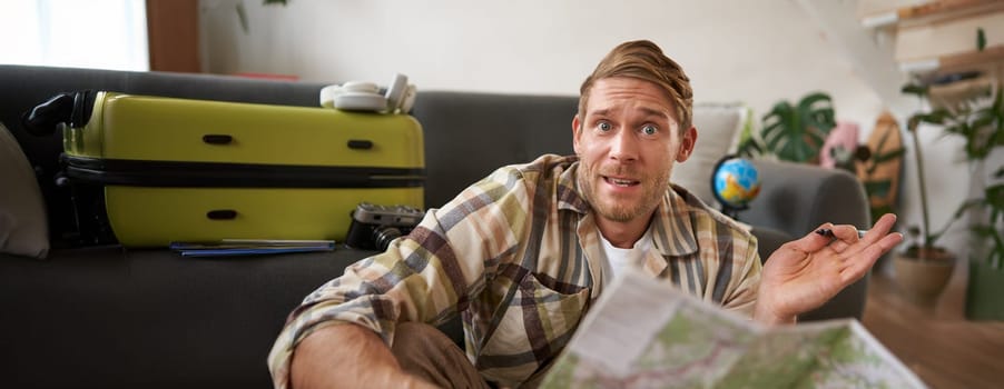 Portrait of man, tourist looking confused at travel map, shrugging, cant decide on route, choosing the road for travelling. Tourism concept