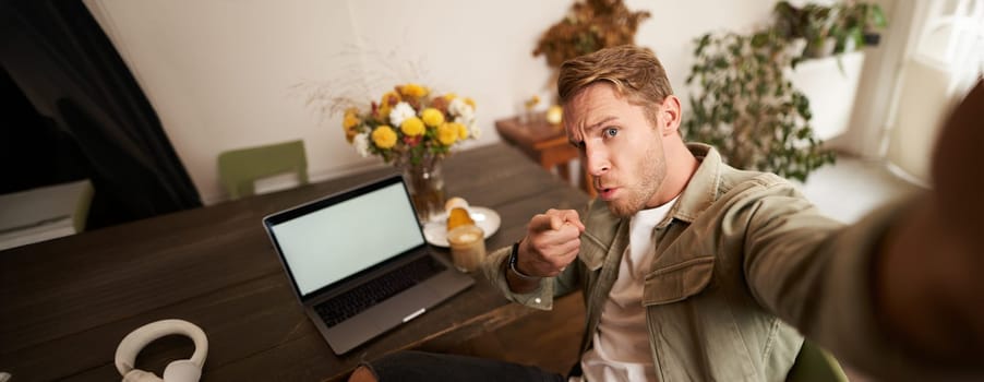 Portrait of serious man takes selfie with fist and threatening face, sits in cafe, poses in coffee shop in front of table with laptop and glass of coffee.
