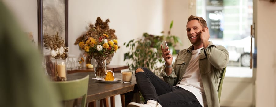 Portrait of handsome, stylish young man, cafe visitor, sitting with his smartphone and listening to music in wireless earphones, smiling at camera.
