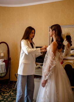 A stylist or makeup artist prepares a bride for a wedding indoors. The background is a warm-toned interior.