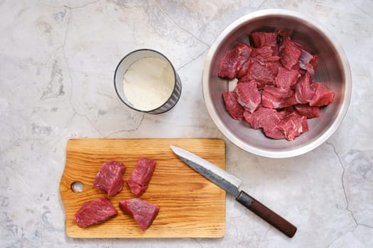 Chunks of fresh beef on a wooden cutting board alongside a sharp knife. A stainless steel bowl filled with additional beef pieces and a tin with flour nearby.