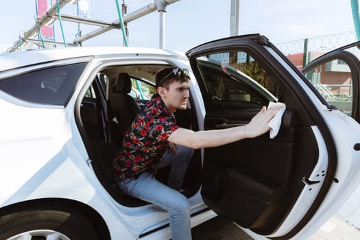 Full-length portrait of a handsome young Caucasian man carefully wiping the black leather upholstery of the rear passenger door of a white car with a damp cloth on a sunny spring day at a city car wash, side view, general plan. Concept: car wash time, working people.