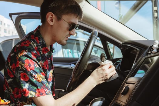Portrait of one young Caucasian handsome brunette man in sunglasses, who is sitting sideways and wiping the black side panel of the steering wheel of a car with a wet napkin on a sunny spring day at a city car wash, side view close-up. Concept time to wash a car, working people.